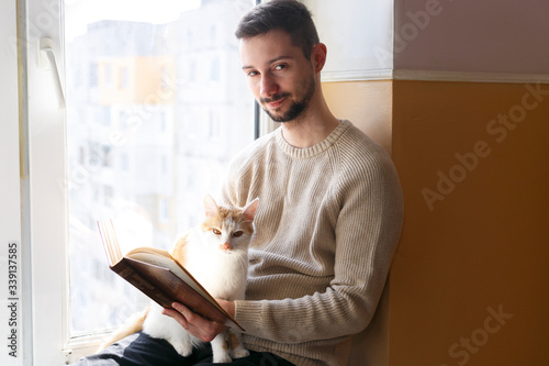 A young man sits on a windowsill and reads a book. Next to the man sits a white cat with red spots. A man is wearing a beige sweater.