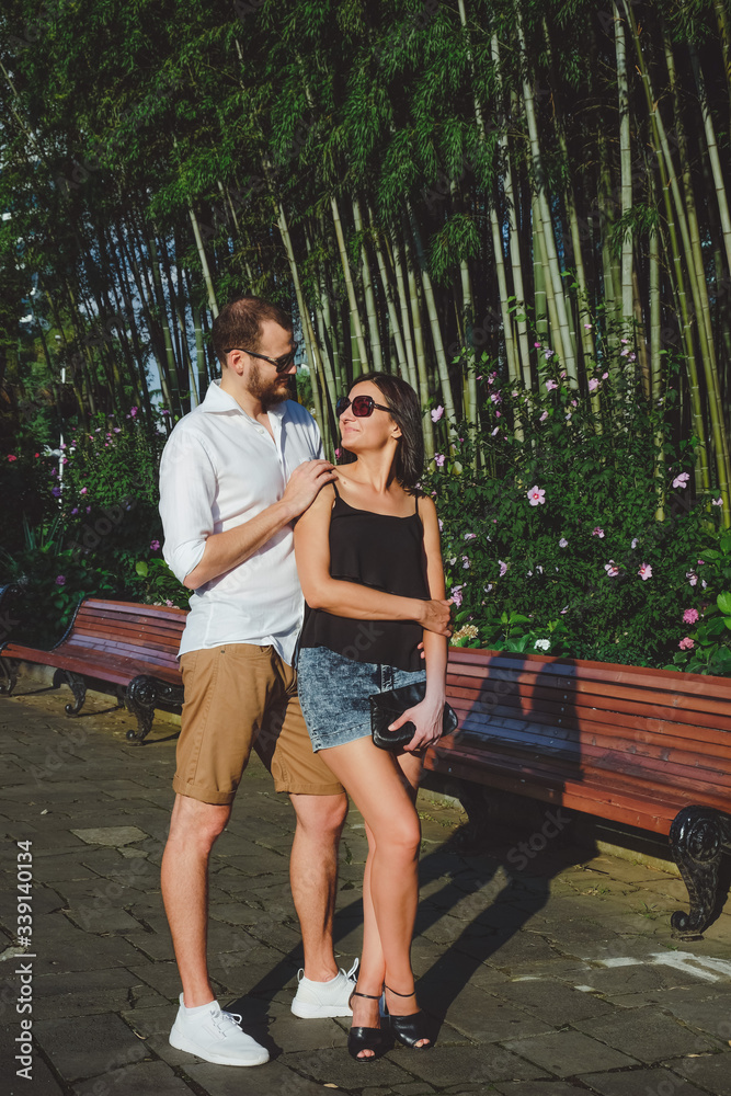 Happy couple of lovers walking outdoors. Bamboo gardens at background. Cheerful, smiling man and lucky lady in sunglasses have fun time in summer. Happy couple of lovers walking outdoors. Bamboo gardens at background. Cheerful, smiling man and lucky lady in sunglasses have fun time in summer.