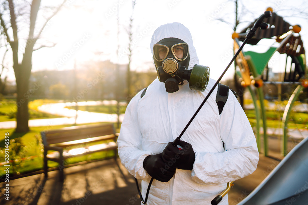 Man wearing protective suit disinfecting the playground in the sun with spray chemicals to preventing the spread of coronavirus, pandemic in quarantine city. Covid -19. Cleaning concept. Man wearing protective suit disinfecting the playground in the sun with spray chemicals to preventing the spread of coronavirus, pandemic in quarantine city. Covid -19. Cleaning concept.