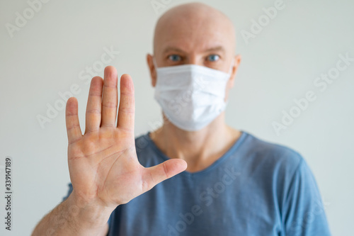 A bald man holds a medical mask in his hands, his other hand held out in protest