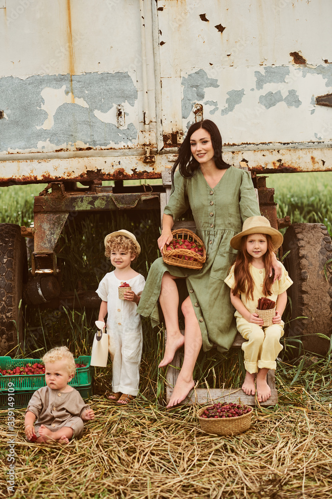 Beautiful young caucasian mother with her children in a linen dress with a basket of strawberries gathers a new crop and has fun with the children near the trailer