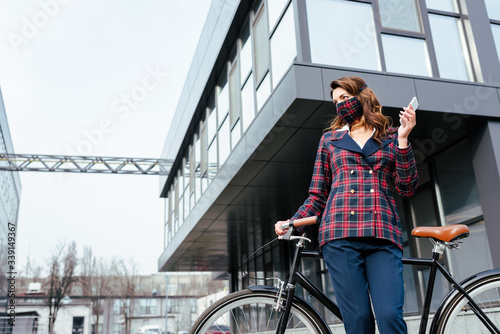 businesswoman in plaid mask standing near bicycle with smartphone