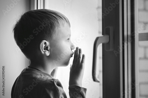 The kid looks out the window during quarantine Covid-19 at home. A boy near the window. The child is on self-isolation at home to protect against coronavirus. Stay at home, let's be healthy.