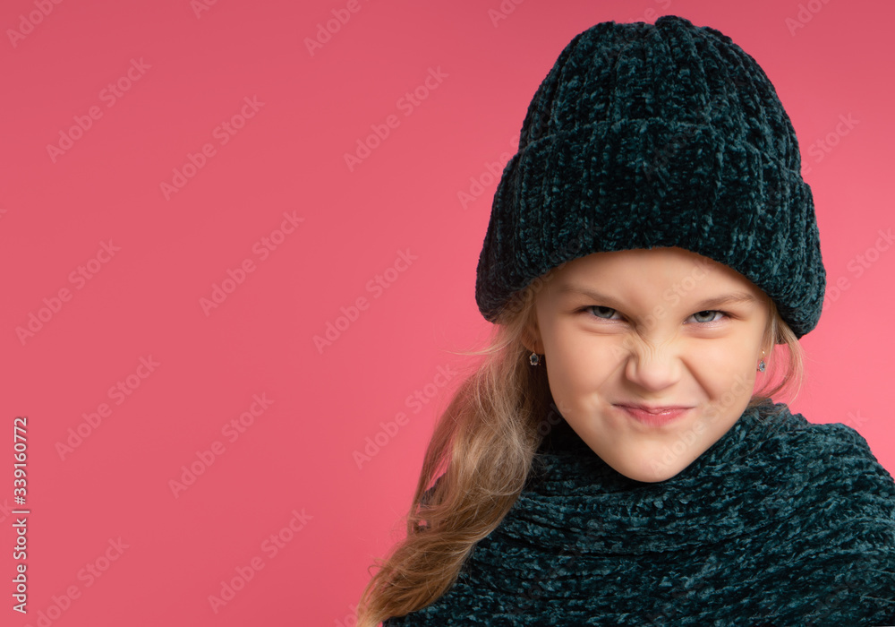 Little blonde kid dressed in green warm scarf and hat. She is smiling, looking naughty, posing against pink studio background. Childhood, fashion, advertising concept. Close up, copy space Little blonde kid dressed in green warm scarf and hat. She is smiling, looking naughty, posing against pink studio background. Childhood, fashion, advertising concept. Close up, copy space