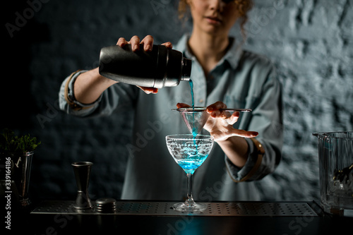 Professional female bartender pours finished blue cocktail to glass.