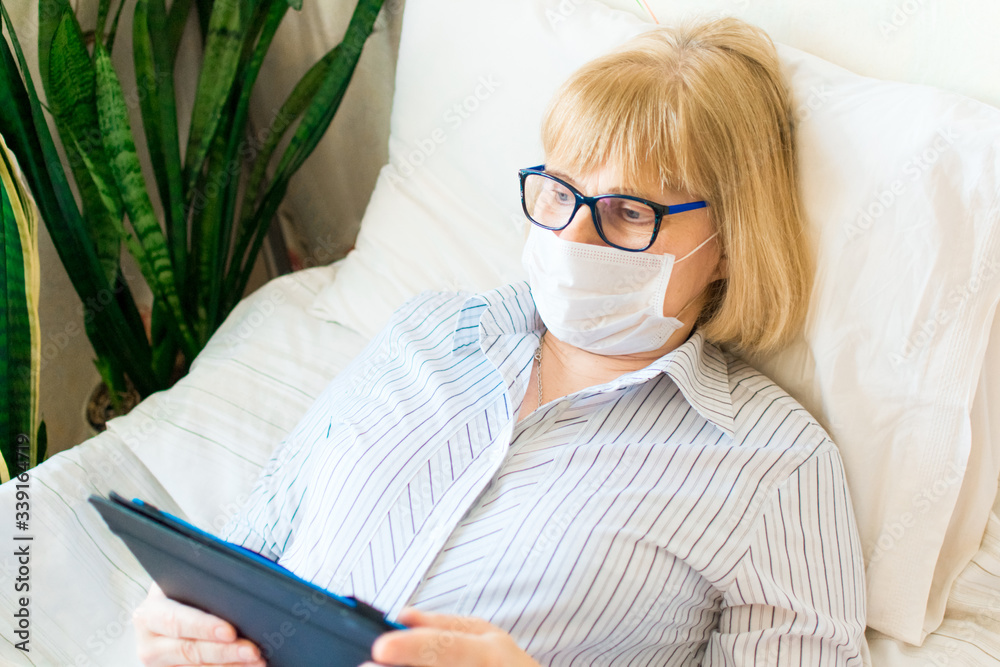 Portrait of adult serious blonde woman in glasses and face mask using tablet lying on bed at home. Remote learning. Social distancing protect coronavirus or covid-19. Online education and work concept