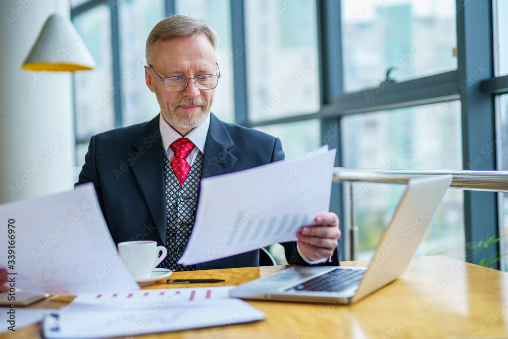 Thoughtful middle aged businessman in suit with a laptop sitting near the window working with documents, typing. Thoughtful middle aged businessman in suit with a laptop sitting near the window working with documents, typing.