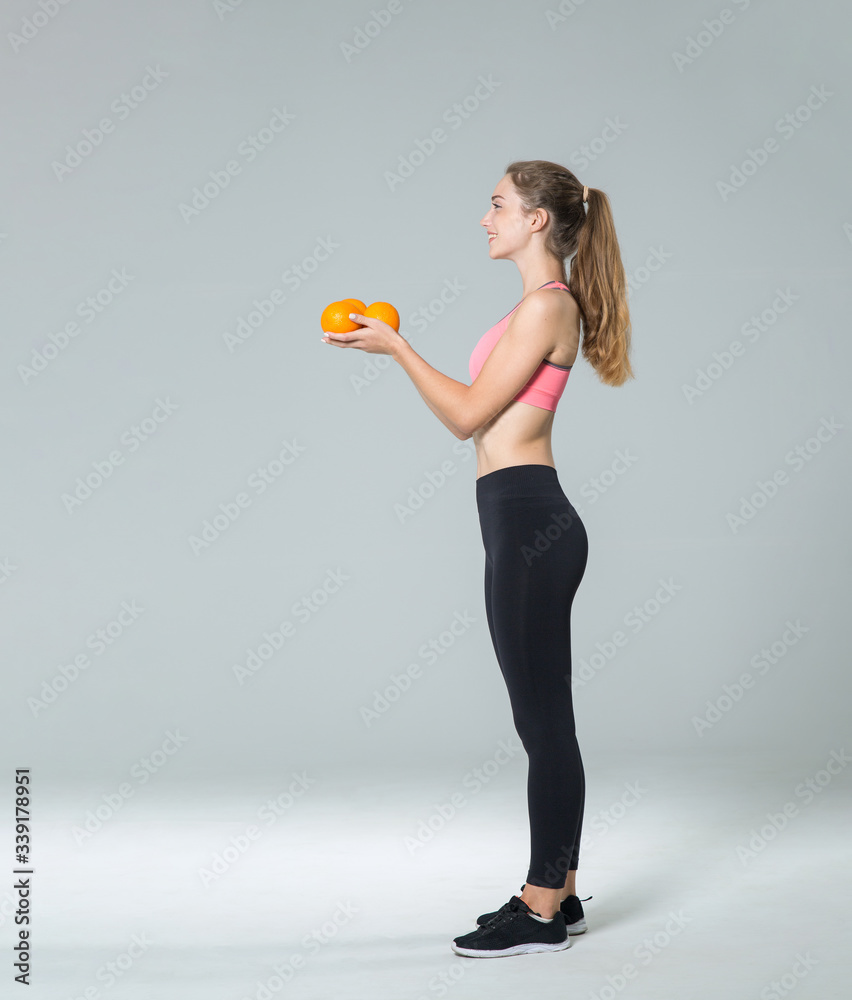 Full height young fitness woman with citrus oranges on gray background. Side view. Full height young fitness woman with citrus oranges on gray background. Side view.