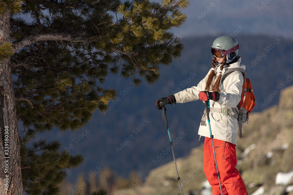 a skier in a bright ski suit stands by a large cedar against the background of a mountain valley with a collapse of large stones. Girl On the Ski. Winter fun at the ski resort. the piste in Alps