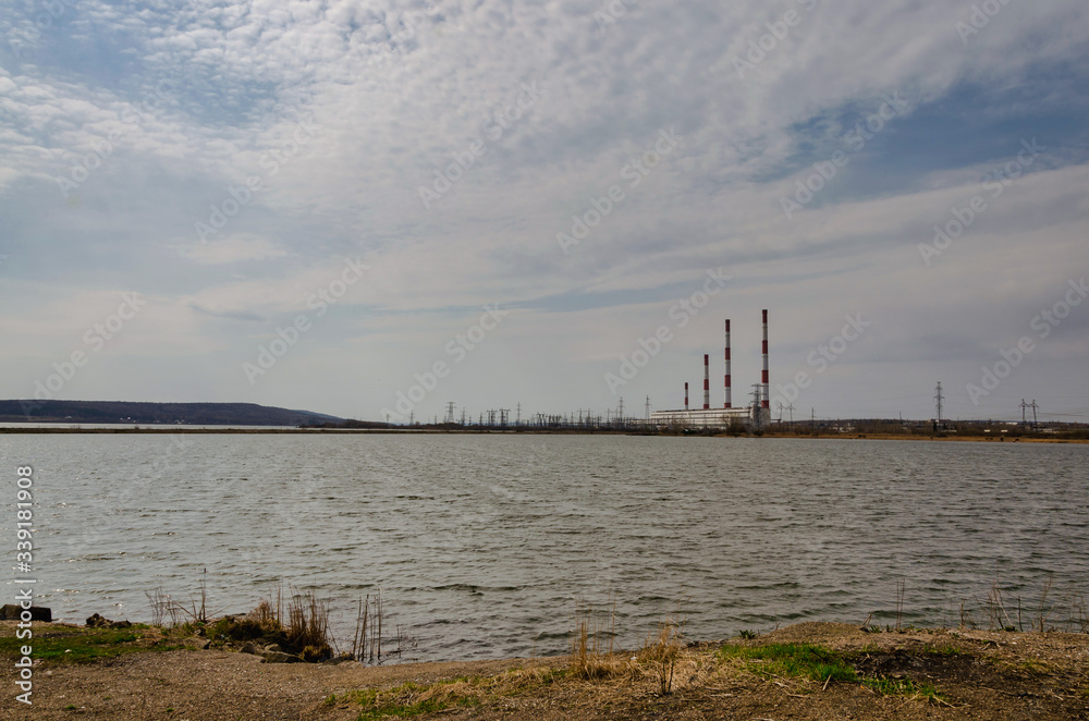 Landscape, four pipes of a power plant on the shore of a reservoir in a spring cloudy day, against a blue sky with clouds.