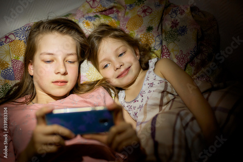 two sisters with a smartphone at night on a pillow, children watch cartoons at night on a smartphone