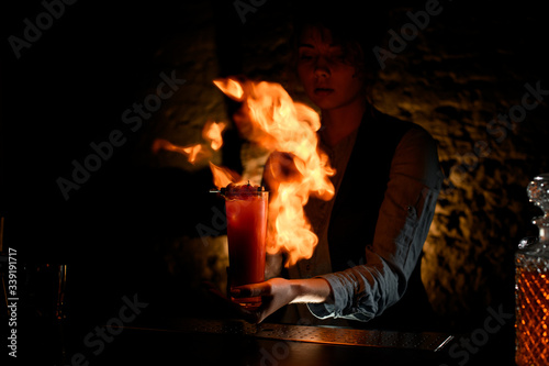 Woman bartender holding glass with cocktail sprinkles on it and masterfully make fire.