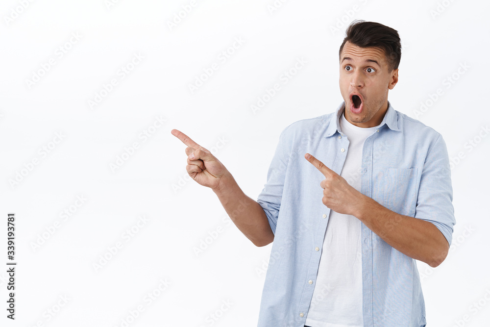 Amazed and shocked young handsome man staring at awesome new product in stock, pointing fingers left and looking with opened mouth at advertisement, standing white background Amazed and shocked young handsome man staring at awesome new product in stock, pointing fingers left and looking with opened mouth at advertisement, standing white background