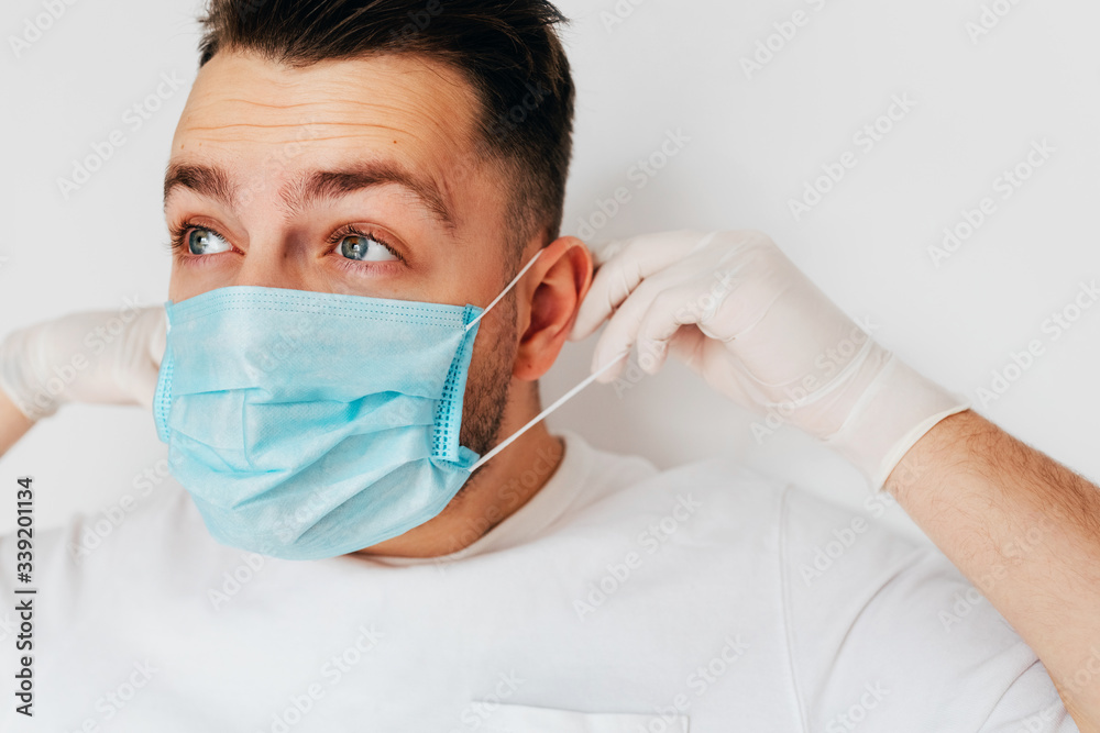 A young man in protective gloves putting on a medical blue mask on light background. Block and quarantine to fight infection Corona virus A young man in protective gloves putting on a medical blue mask on light background. Block and quarantine to fight infection Corona virus