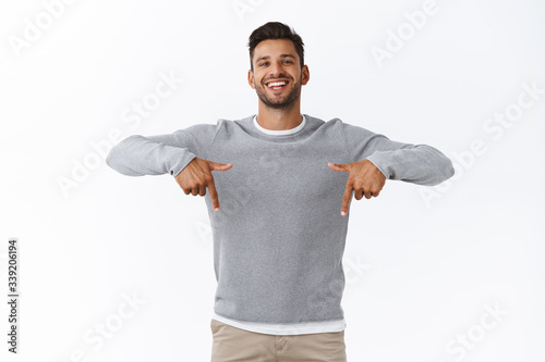 Happy carefree, friendly handsome man with bristle in grey casual sweater, joyfully talking, give advice, recommend promo as pointing bottom, indicate down link, standing white background
