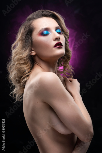 Beautiful topless busty girl with blue makeup poses on black and pink background, covering her breast with her hands. Fashionable, advertising and commercial portrait design.