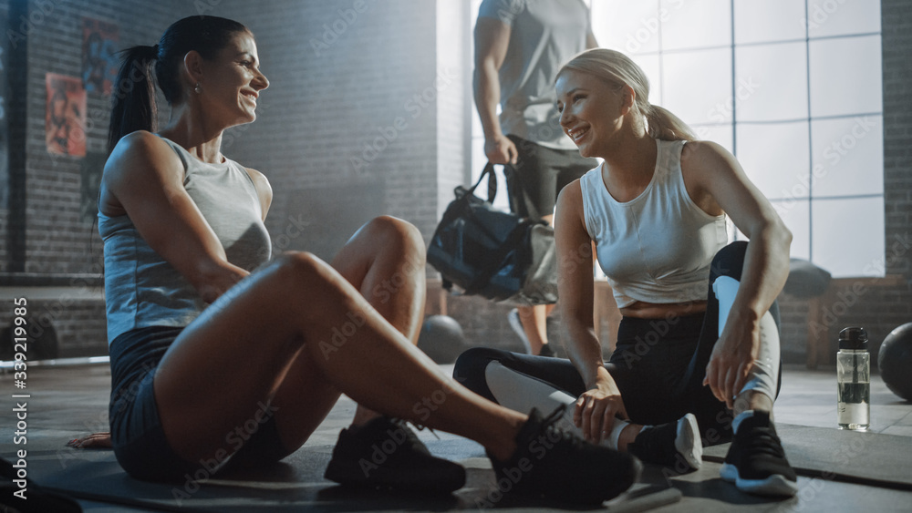 Two Beautiful Fit Athletic Girls Sit on a Floor of Industrial Loft Gym. They're Happy with their Training Program and Successfully Give a High Five. Strong Masculine Man Walks in the Background. Two Beautiful Fit Athletic Girls Sit on a Floor of Industrial Loft Gym. They're Happy with their Training Program and Successfully Give a High Five. Strong Masculine Man Walks in the Background.