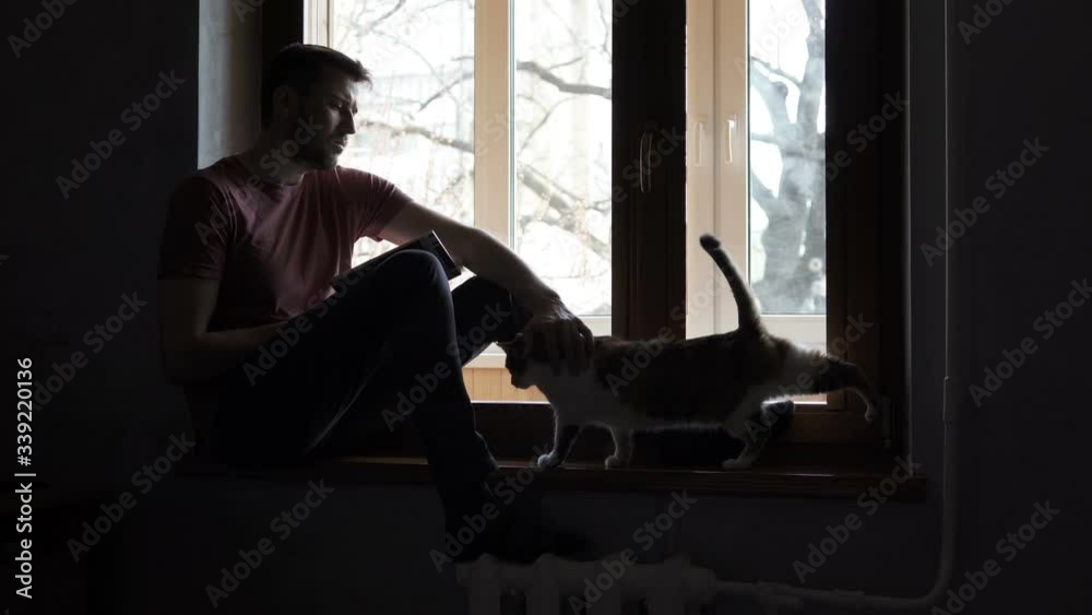 An attractive guy is reading a book sitting on a windowsill in a house with a charming cat. Self-isolation, quarantine, pandemic concept. An attractive guy is reading a book sitting on a windowsill in a house with a charming cat. Self-isolation, quarantine, pandemic concept.
