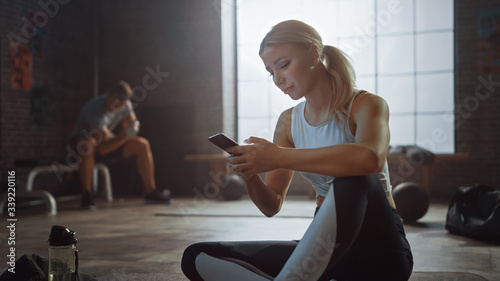 Happy and Smiling Beautiful Athletic Young Woman is Using a Smartphone while Sitting on a Floor in a Loft Gym. She's Typing a Message and Smiling. A Man Exercises in the Background. 