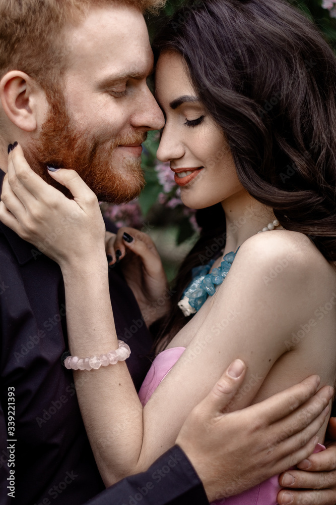 Beautiful couple in love cuddling in a blooming lilac garden. A woman in a purple sleeveless dress with a delicate necklace around her neck. Red-haired man in a black suit kisses and hugs a girl