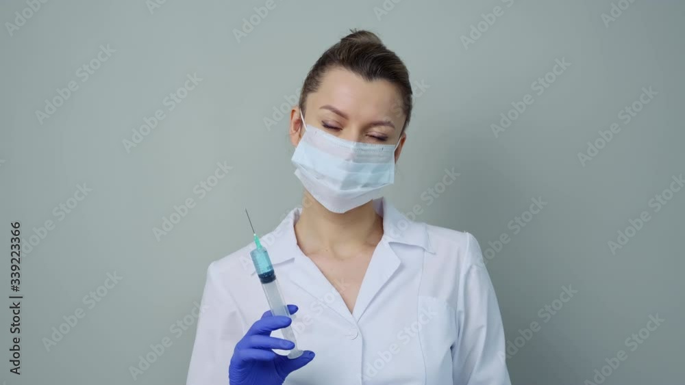 A female medical worker in a white robe and mask holds a syringe with a prick of liquid. A female medical worker in a white robe and mask holds a syringe with a prick of liquid.