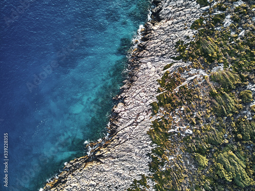 top drone view of a stony coast clashing into the blue ocean sea. Structure and Pattern in artistic way of the sharp rocks in the water. Aerial top shot on warm summer waves.