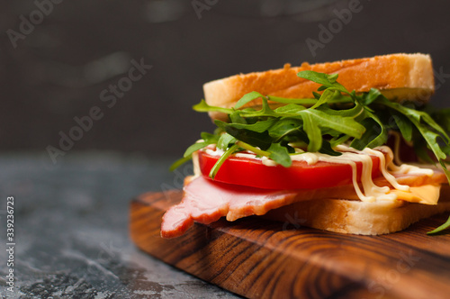 Very tasty big sandwich with ham or balyk, arugula, cheese, tomatoes and mayonnaise on a cutting board in rustic style on a gray background view from the side, copy space