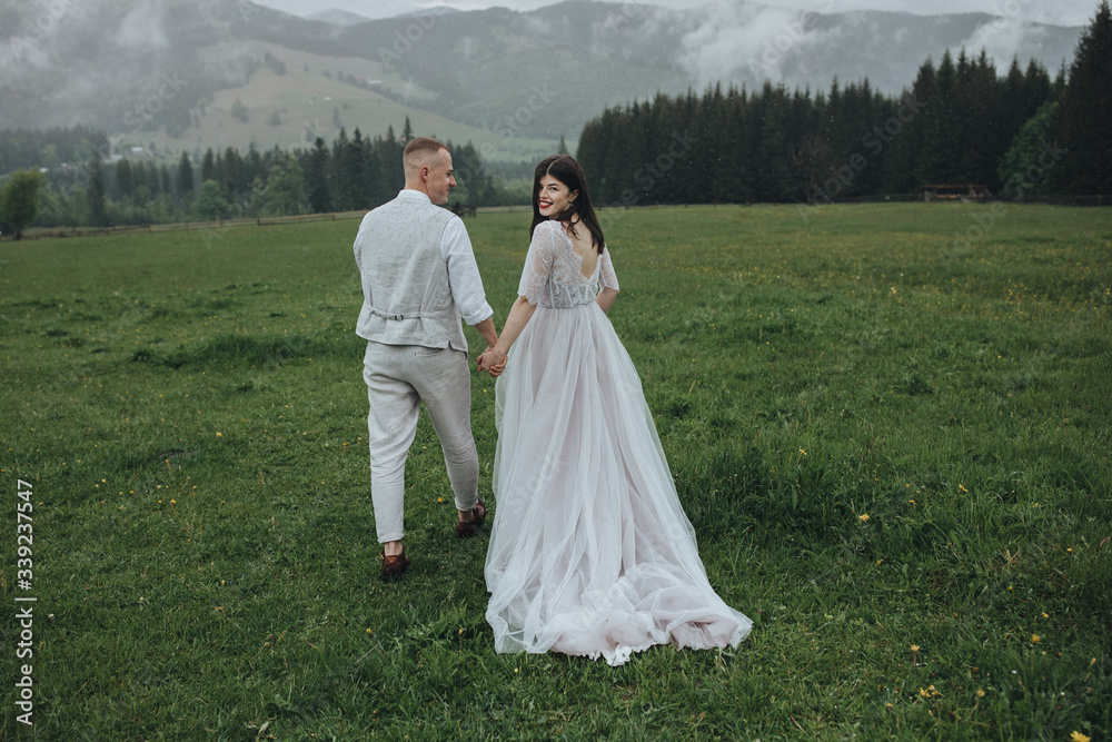 Spring wedding in the mountains. A guy in a shirt and vest and a girl in a white dress walk along a wooden fence along a green meadow against the backdrop of mountains and forests