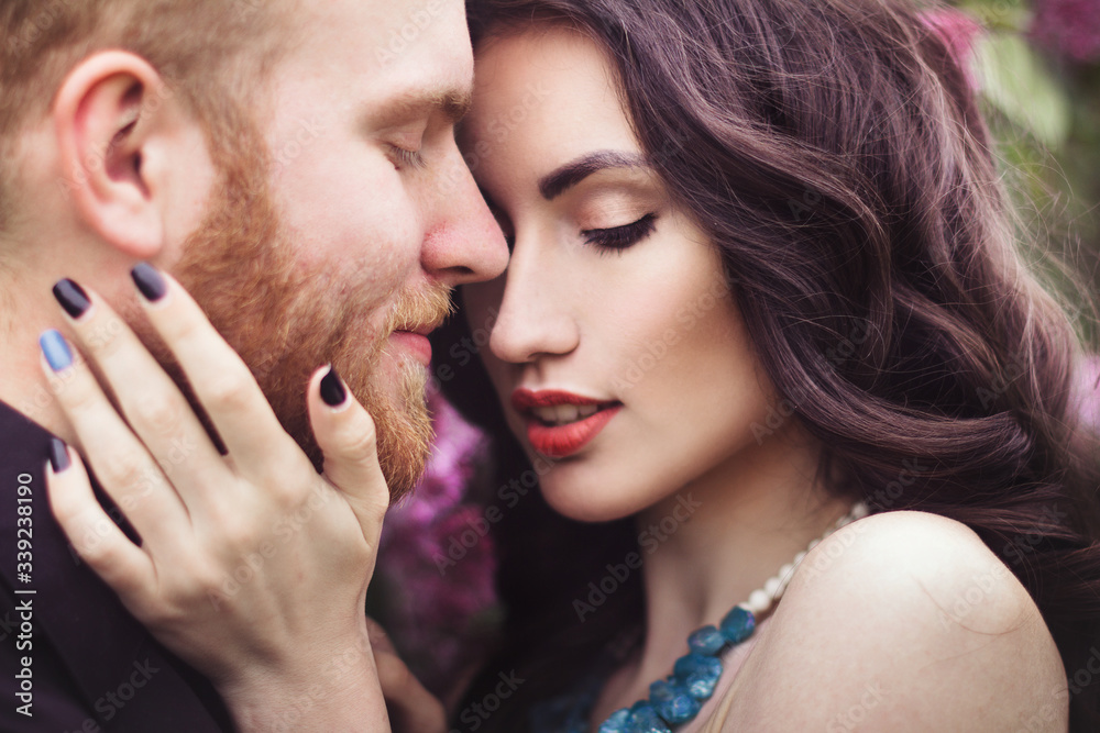 Beautiful couple in love cuddling in a blooming lilac garden. A woman in a purple sleeveless dress with a delicate necklace around her neck. Red-haired man in a black suit kisses and hugs a girl Beautiful couple in love cuddling in a blooming lilac garden. A woman in a purple sleeveless dress with a delicate necklace around her neck. Red-haired man in a black suit kisses and hugs a girl
