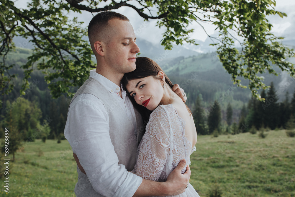 Spring wedding in the mountains. A young guy in a white shirt and trousers and a girl in a white dress are standing under the branches of a large tree in the mountains at sunset Spring wedding in the mountains. A young guy in a white shirt and trousers and a girl in a white dress are standing under the branches of a large tree in the mountains at sunset
