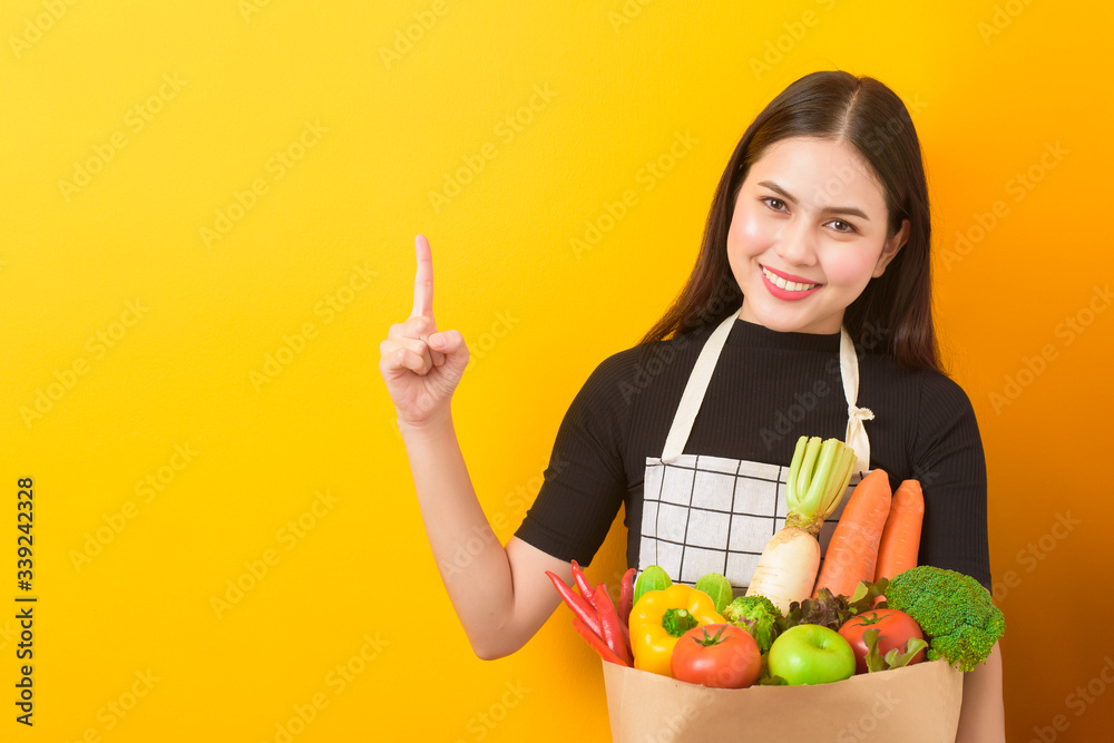 Beautiful young woman is holding vegetables in grocery bag in studio yellow background Beautiful young woman is holding vegetables in grocery bag in studio yellow background