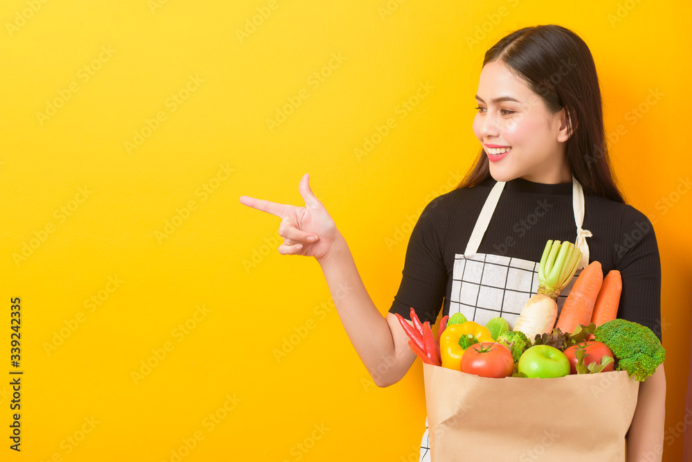 Beautiful young woman is holding vegetables in grocery bag in studio yellow background Beautiful young woman is holding vegetables in grocery bag in studio yellow background