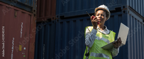 Black foreman woman worker working checking at Container cargo harbor holding laptop and radio to loading containers. African dock female staff business Logistics import export shipping concept.