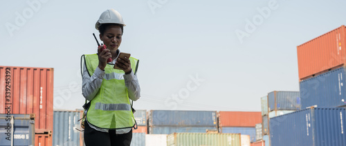 Black foreman woman worker working checking at Container cargo harbor holding radio and smartphone to loading containers. African dock female staff business Logistics import export shipping concept.