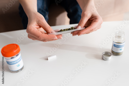 cropped view of mature woman holding paper with dried weed near bottles with medical cannabis lettering