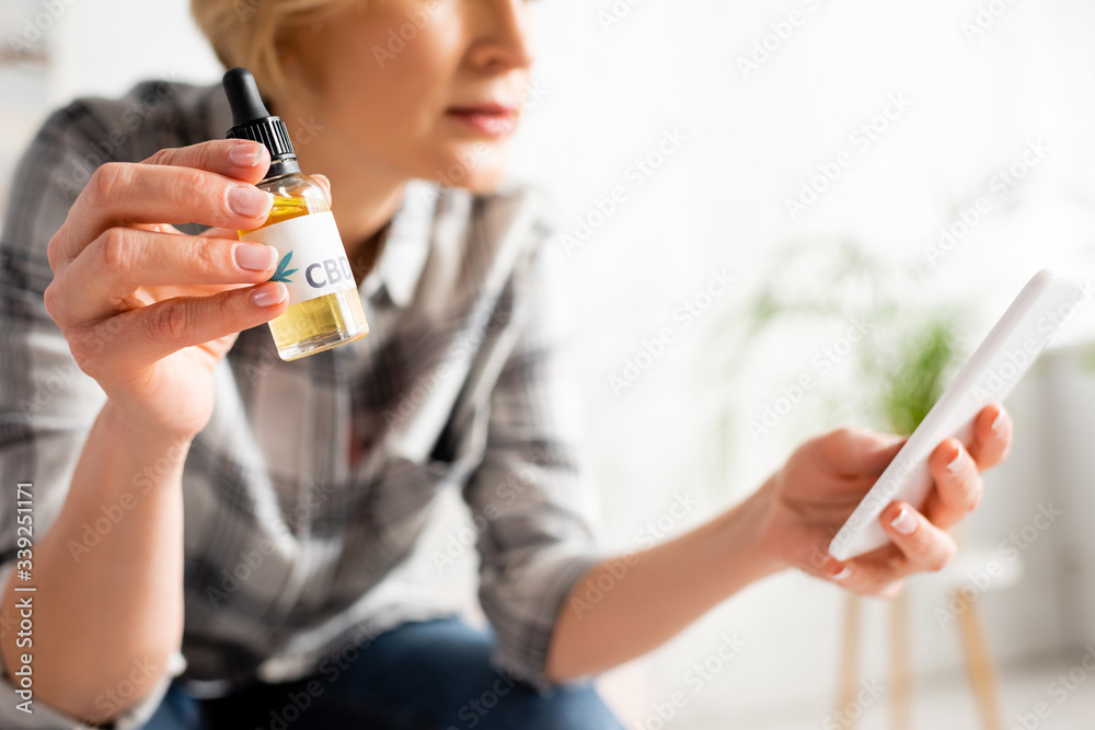 cropped view of mature woman holding bottle with cbd lettering and using smartphone at home