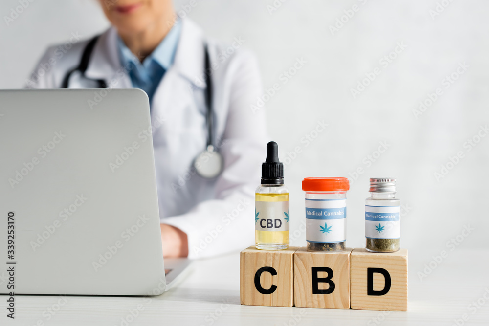 cropped view of doctor using laptop near cubes with cbd lettering and bottles with medical cannabis cropped view of doctor using laptop near cubes with cbd lettering and bottles with medical cannabis