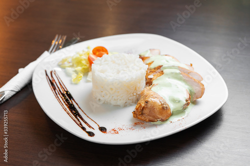Chicken fillet with sauce is served with rice and vegetables on a white plate and a dark table.