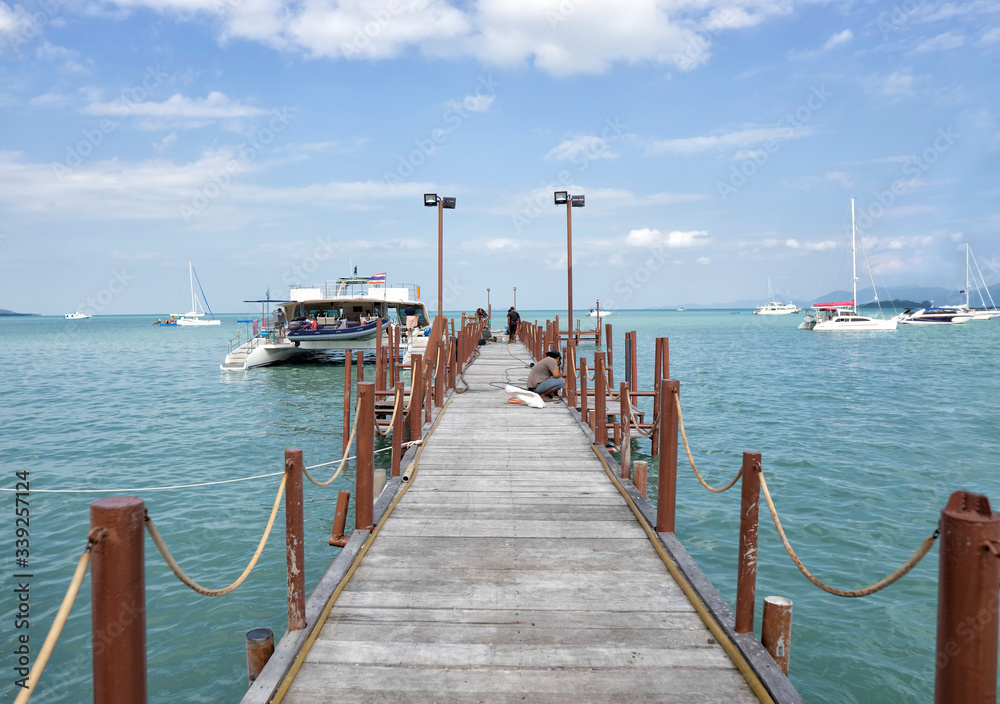 A long wooden pier at the ferry from Koh Samui to Phangan Island in Thailand. A ferry-boat departs from the pier, several yachts float on the horizon on a clear sunny day.