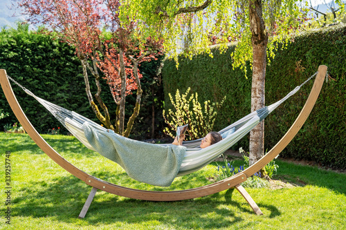 Young relaxed girl reading travel book in hammock in garden at home at bright sunny day. Slow living, gadget detox and weekend leisure activity. Quarantine and self isolation period photo