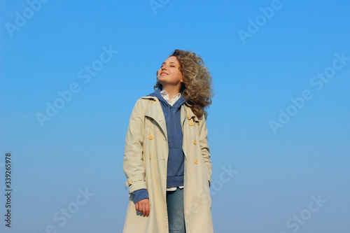 Curly young girl wearing a beige coat, blue jeans and hoodie over blue sky background. People, landscapes, travel concept. © diesel_80