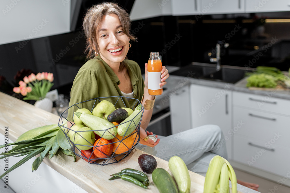 Portrait of a young and cheerful woman drinking juice, sitting with healthy raw food on the kitchen at home. Vegetarianism, wellbeing and healthy lifestyle concept - obrazy, fototapety, plakaty 