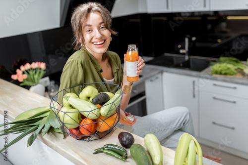Portrait of a young and cheerful woman drinking juice, sitting with healthy raw food on the kitchen at home. Vegetarianism, wellbeing and healthy lifestyle concept