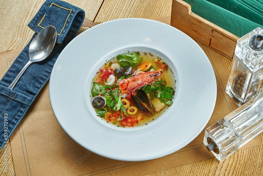 Italian seafood soup with tomato salsa. Bouillon with crevete, mussels and greens in a white plate on a wooden background. Restaurant table setting. Close up