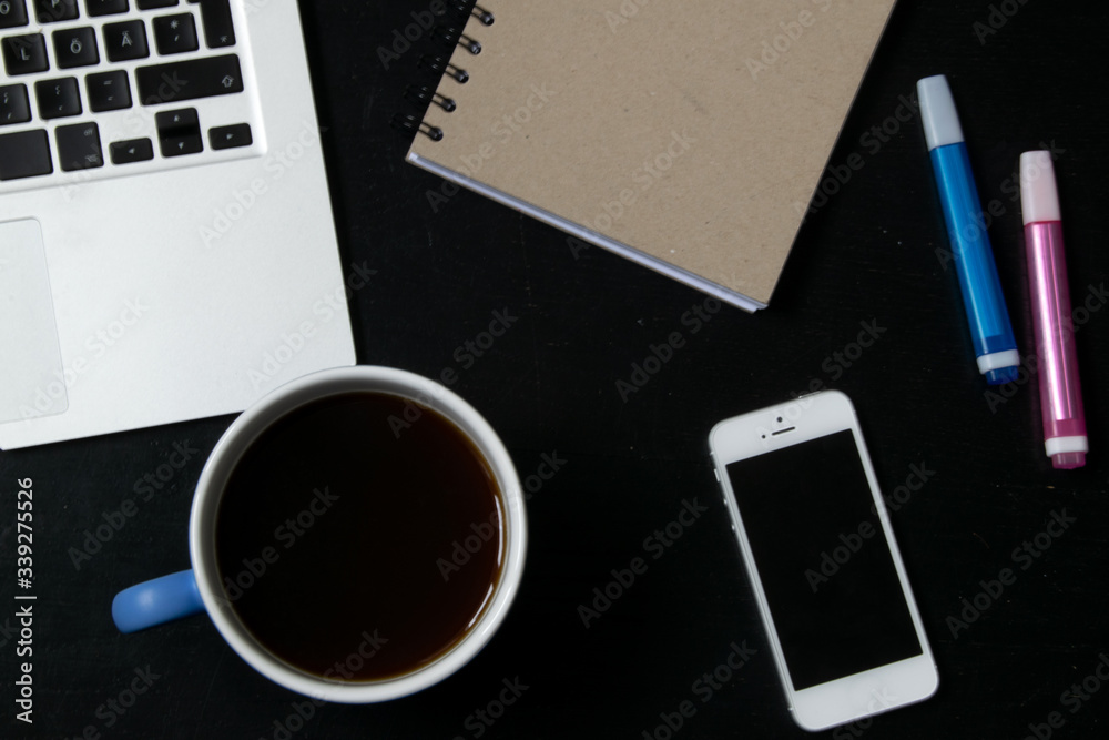 View from above of modern workplace with cup of tea, laptop keyboard, mobile phone, pens and notebook on black wooden table View from above of modern workplace with cup of tea, laptop keyboard, mobile phone, pens and notebook on black wooden table