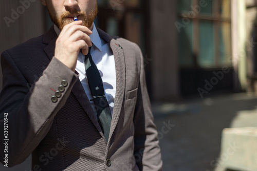 electronic cigarettes iqos. Portrait of a young bearded guy of twenty-five years old, stands by the textured wall of the building, smokes a cigarette. Outdoor photo