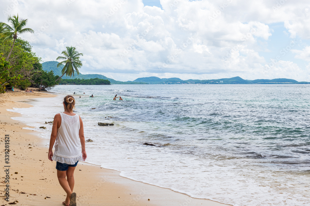 Femme marchant sur une plage de sable blanc avec une mer tropicale bleu d'azur et quelques baigneurs, avec des palmiers sur fond des côtes de Martinique dans un ciel partiellement couvert de nuages Femme marchant sur une plage de sable blanc avec une mer tropicale bleu d'azur et quelques baigneurs, avec des palmiers sur fond des côtes de Martinique dans un ciel partiellement couvert de nuages