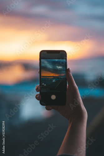 Hand holding a smartphone taking a photograph of the sea at sunset