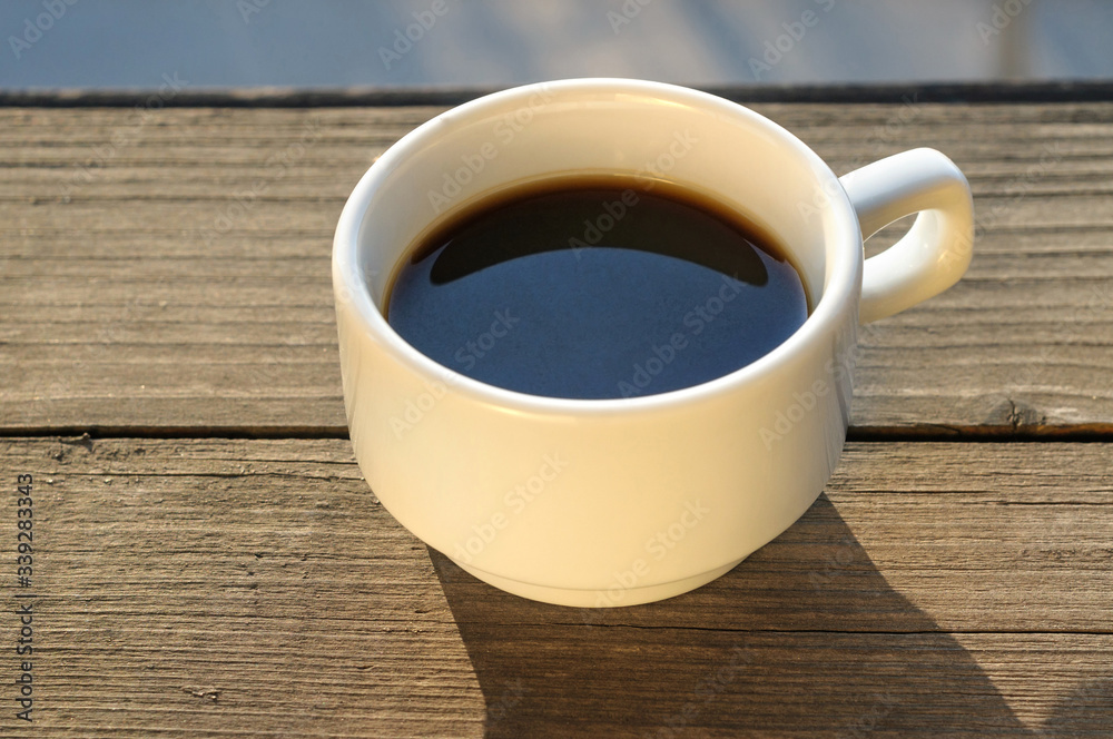 Black coffee in a white cup on a wooden background. White cup with black coffee on the balcony in the rays of the evening sun.