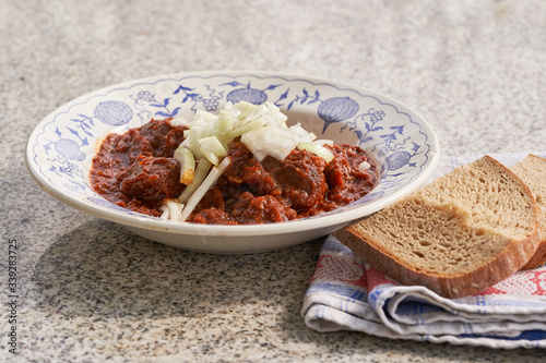 Traditional beef goulash with chopped white onion and slices of rustic bread served in vintage deep plate on the stone table outside. Popular meal in Czech republic, Slovakia, Hungary and Austria.